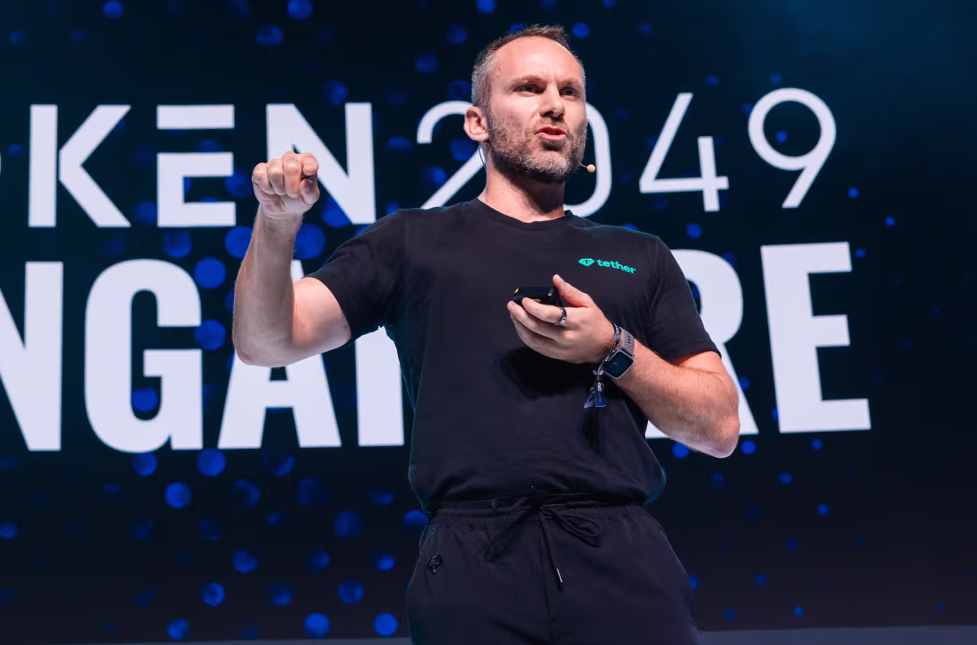 Man in black Tether shirt speaking on stage with a microphone headset and holding a device.